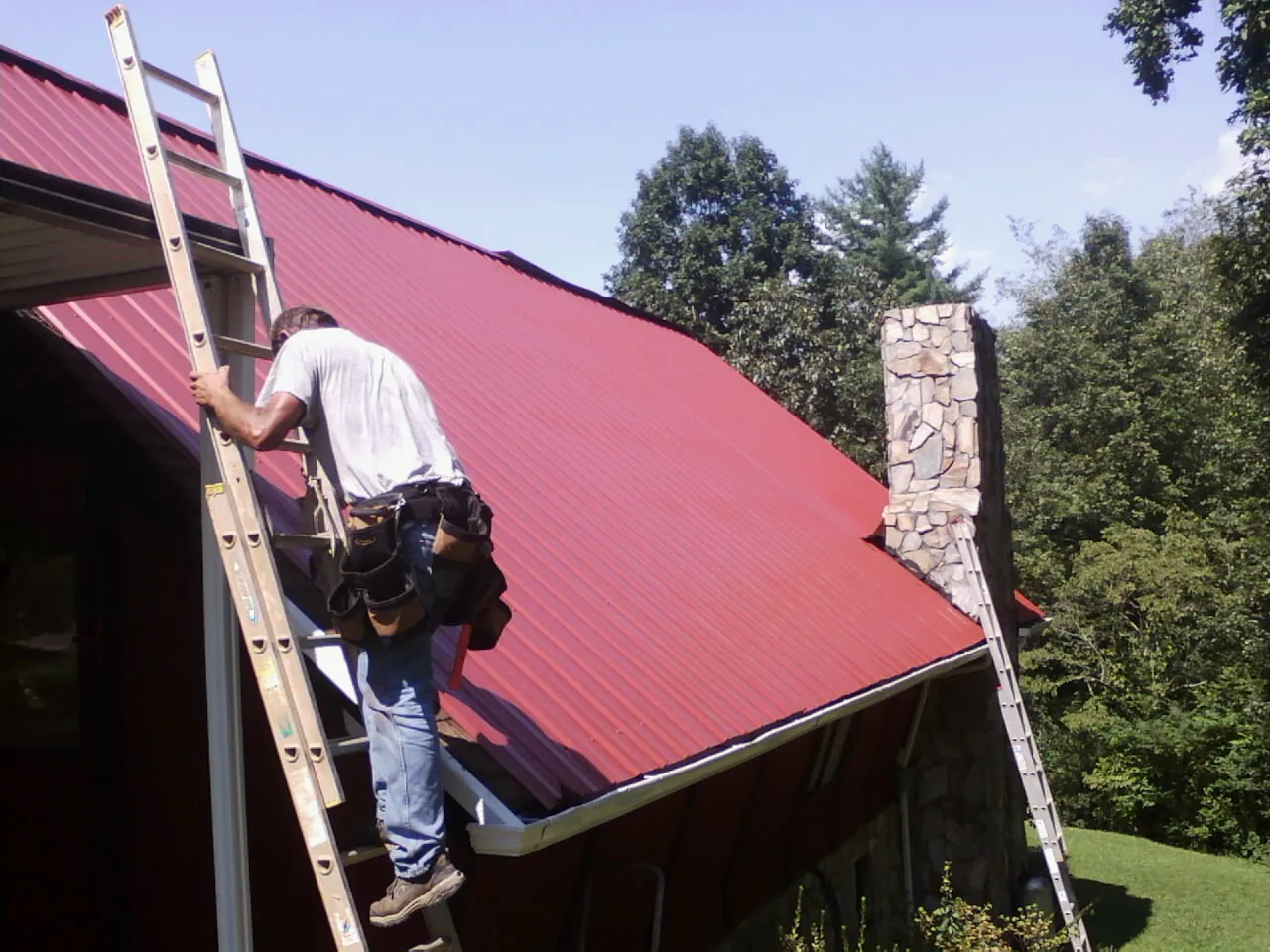 Roofer on a ladder beside a red metal roof and stone chimney — Soffit & Fascia Repair by Roofing Brothers in Peck